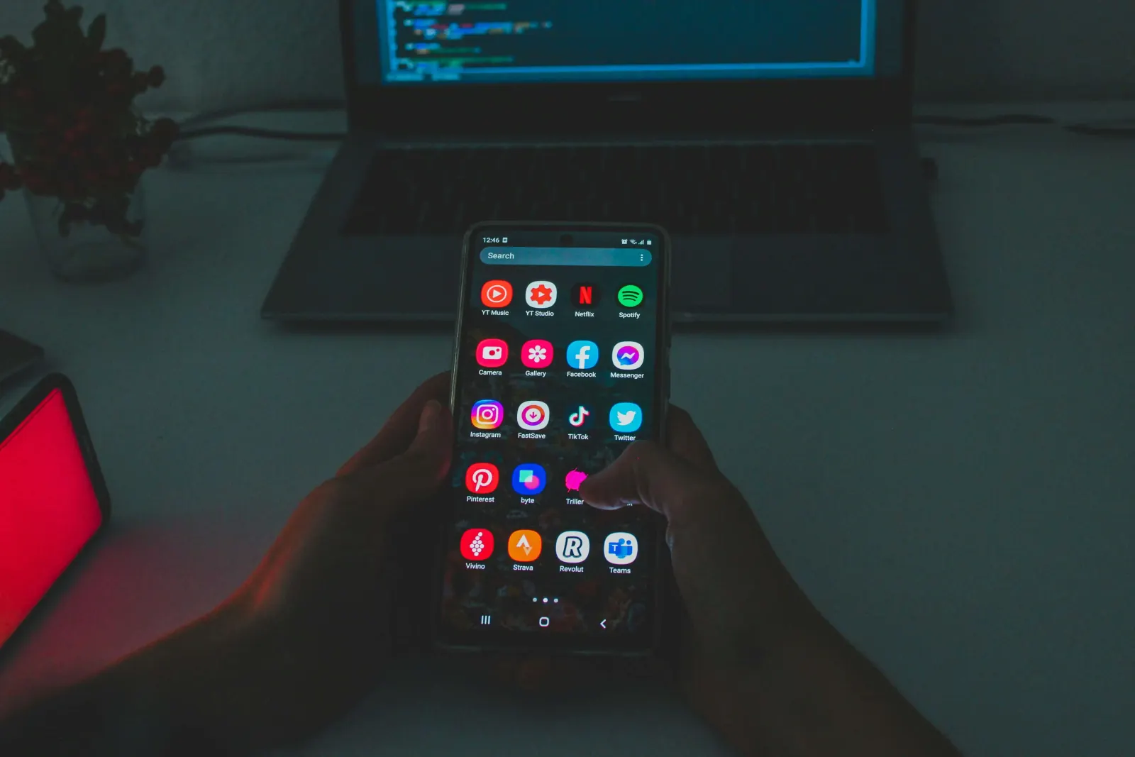 Hand holding a phone with app icons in front of a laptop