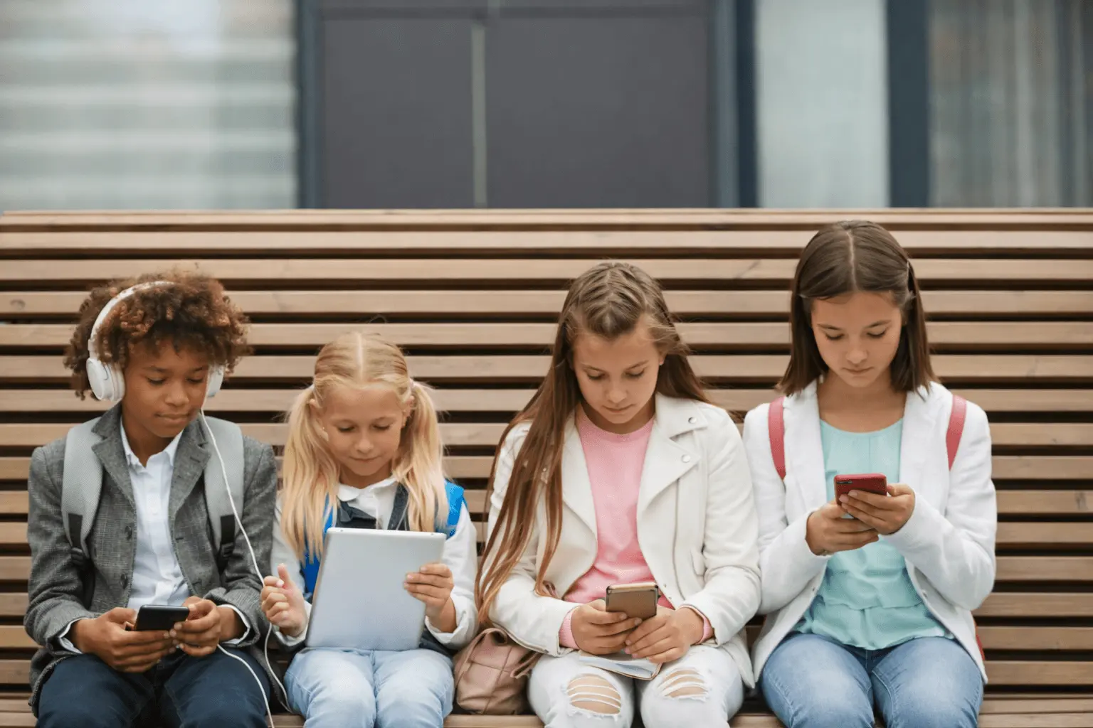Four kids sitting on a bench absorbed in phones and a tablet
