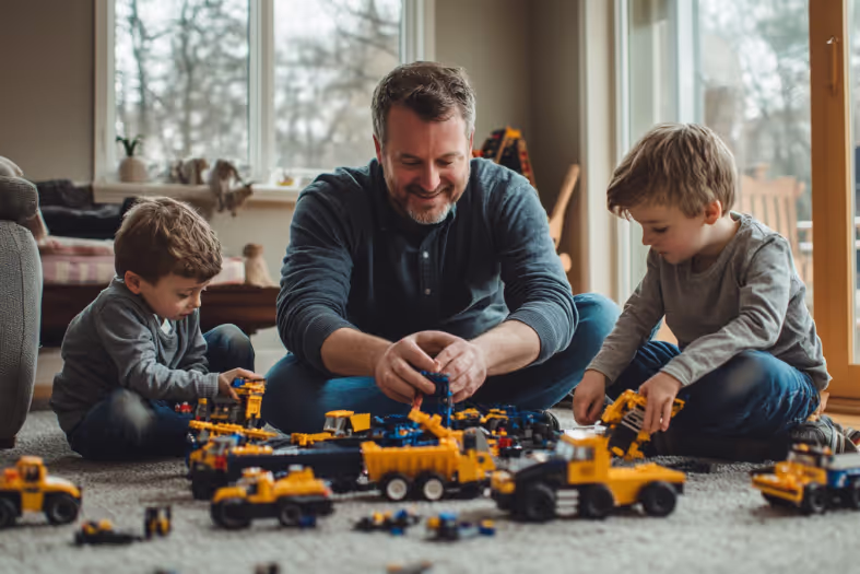 Father and two boys playing with yellow toy construction trucks on the floor