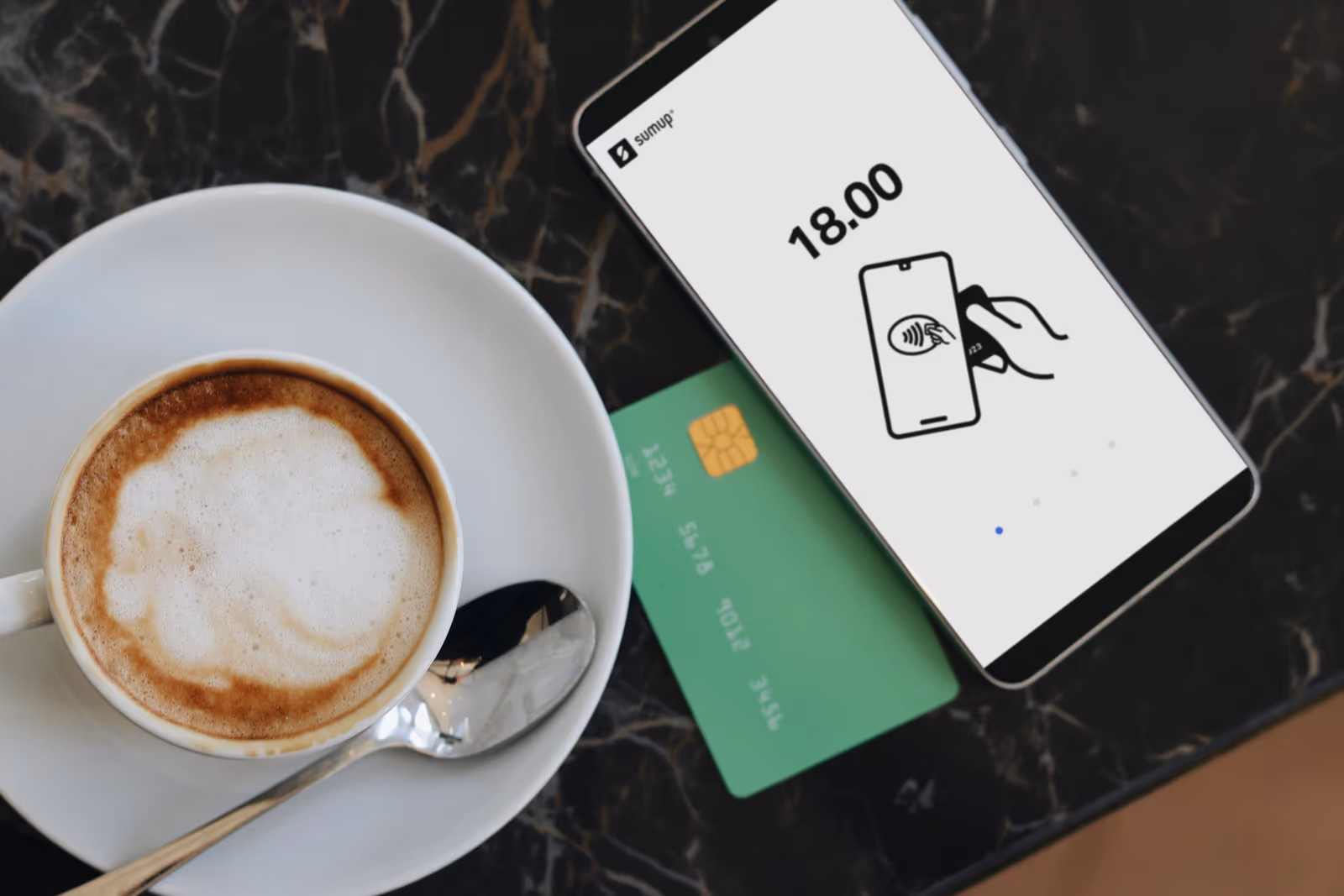 Green payment card beside a phone showing contactless tap-to-pay, next to a cappuccino