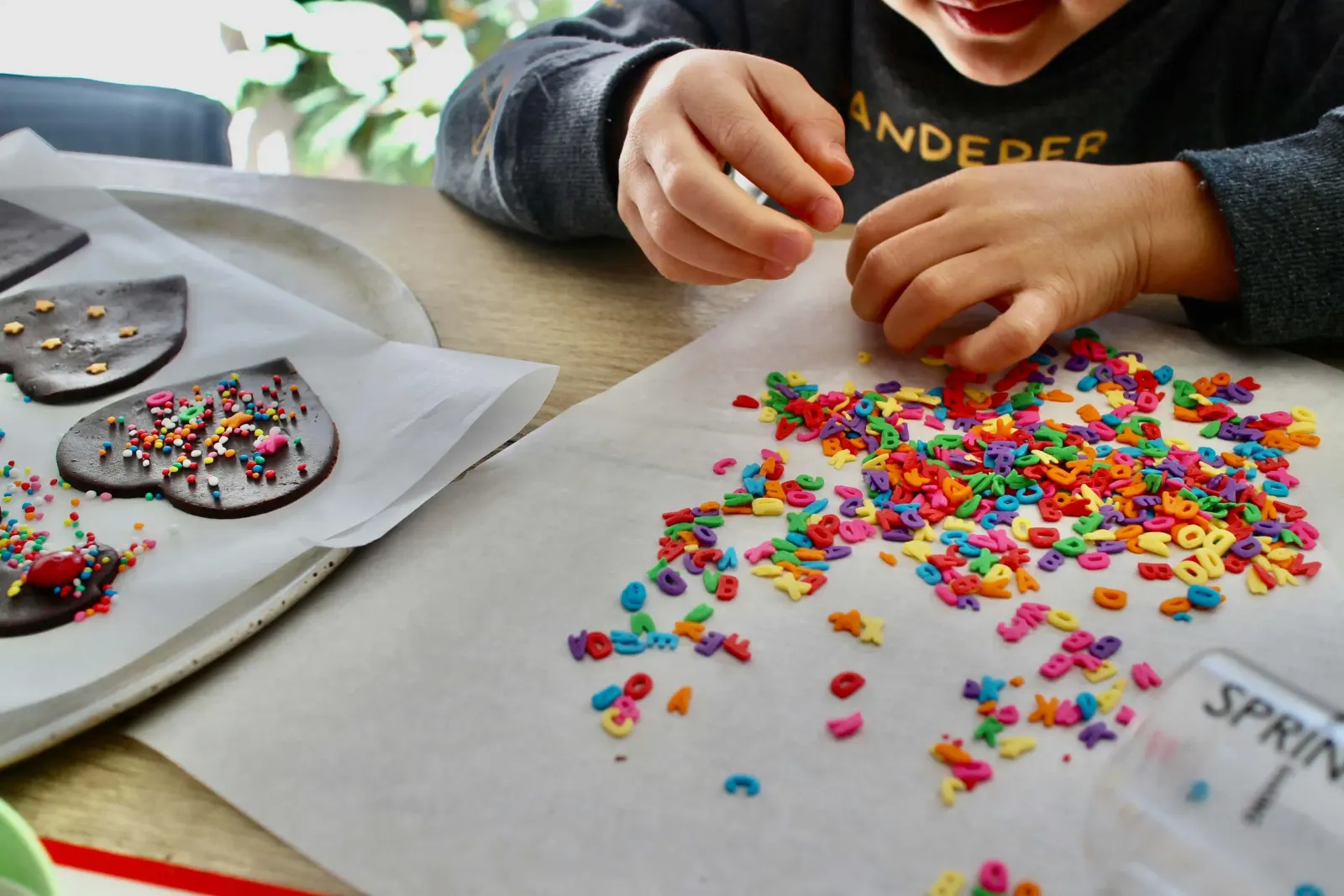Child's hands scattering colorful alphabet sprinkles near frosted heart cookies