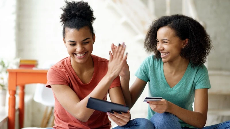 Two young women high-fiving while holding a credit card and phone