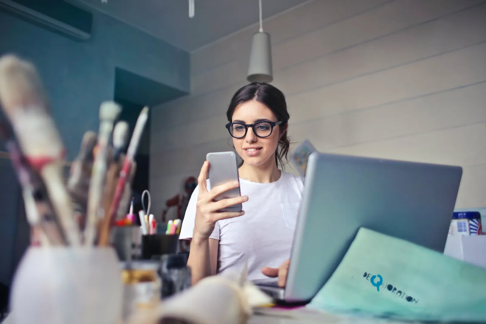 Woman in glasses smiling at her phone beside a laptop