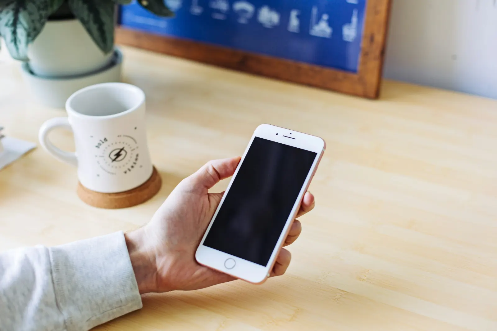 hand holding a rose-gold smartphone with a blank black screen on a desk